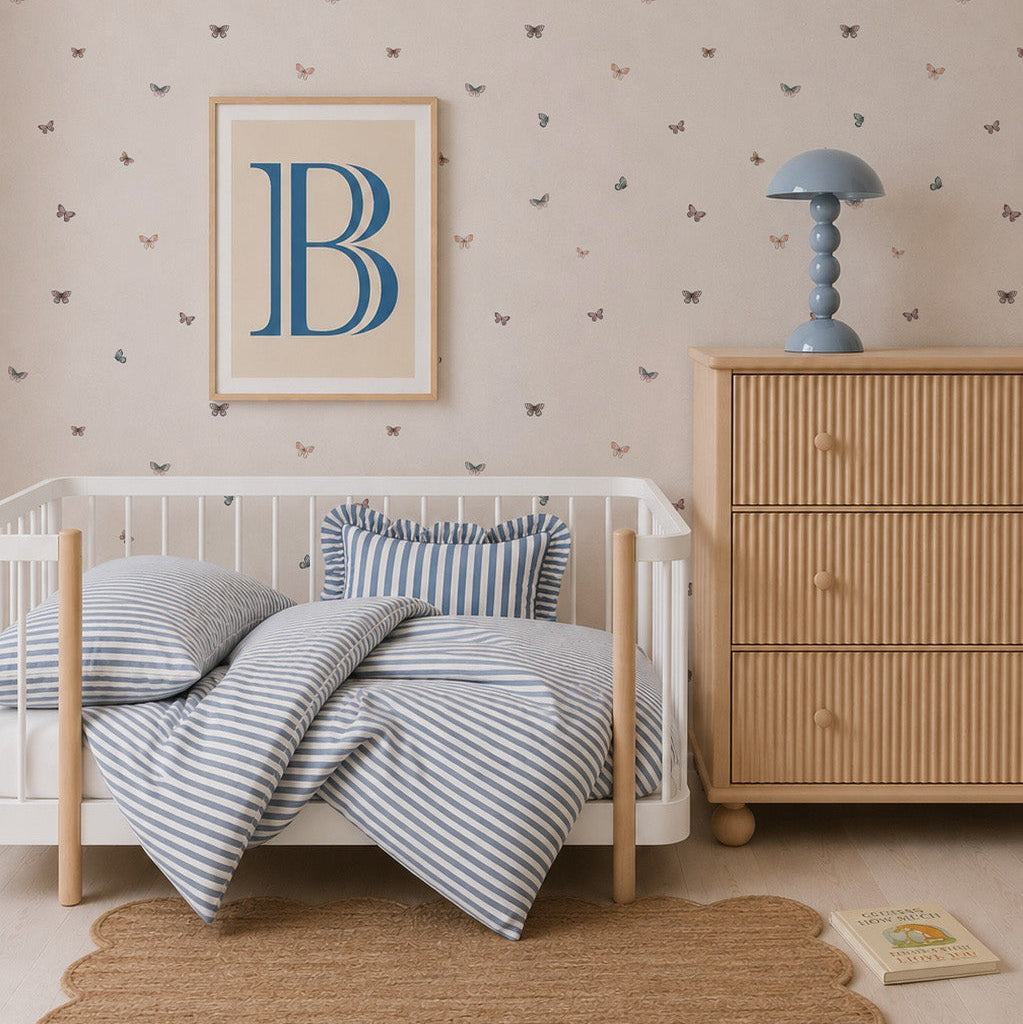 A cozy nursery features a white crib, blue-striped bedding, a wooden dresser, a blue lamp, books scattered on the floor, and charming Bibi Butterflies Wallpaper in a hand-drawn repeat pattern. A framed letter B hangs above the crib.