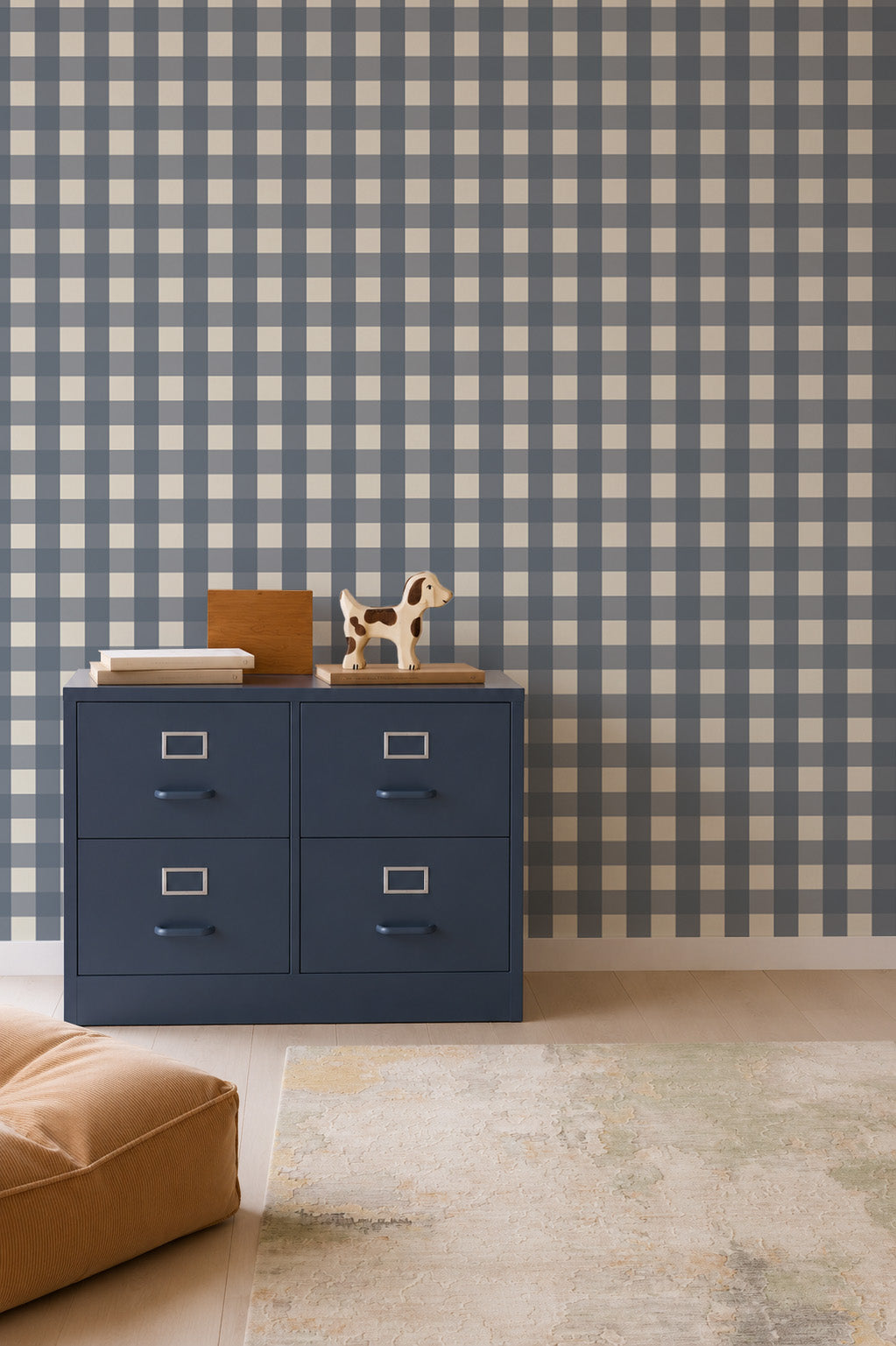A navy blue four-drawer filing cabinet stands against a wall covered in Large Check Wallpaper, Deep Blue, topped with a wooden dog figurine and books.