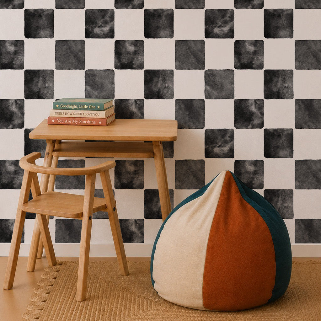 A small wooden desk and chair sit on a woven rug in front of Billys Checkered Wallpaper, Black. Books rest on the desk, a pendant lamp hangs above, and a round color-block beanbag completes this cozy, playful space.