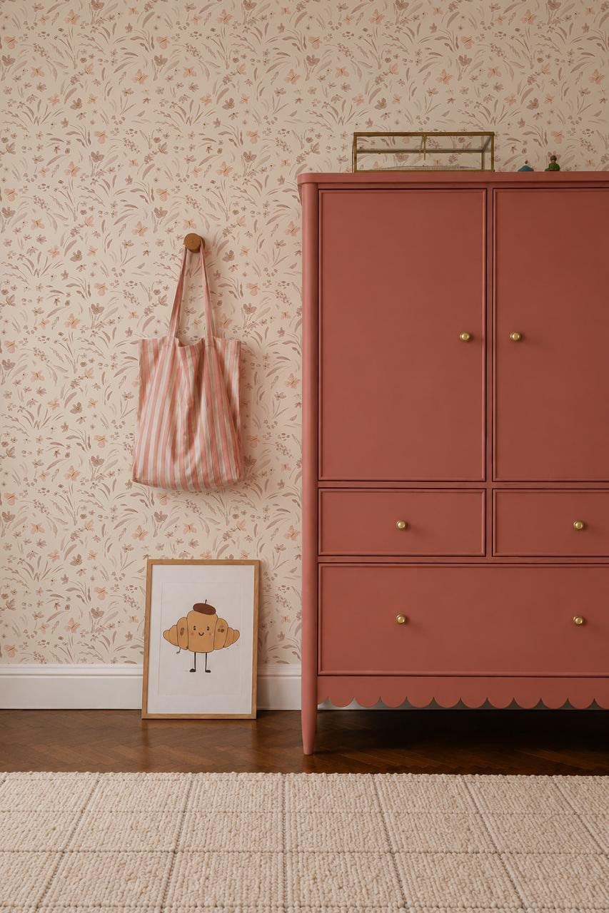 A dresser stands in a room with Butterfly Meadowland Wallpaper, Dune. A pink striped tote hangs on a wood peg, and a framed muffin illustration rests on the wooden floor beside a beige rug.