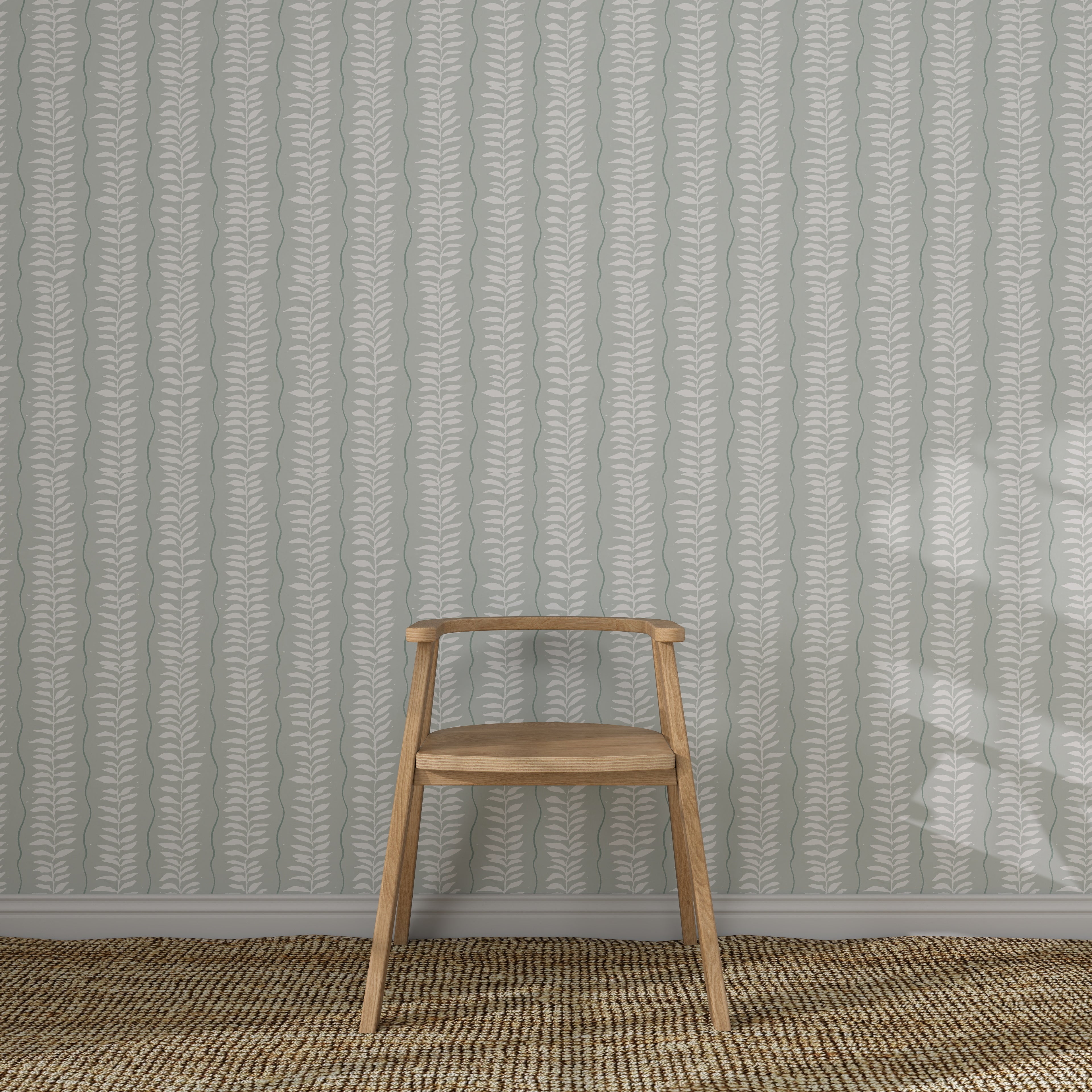A simple wooden chair sits on a woven rug in front of Meadow Leaf Stripe Wallpaper, Sea Foam, featuring hand-painted vertical leaf designs in muted tones.