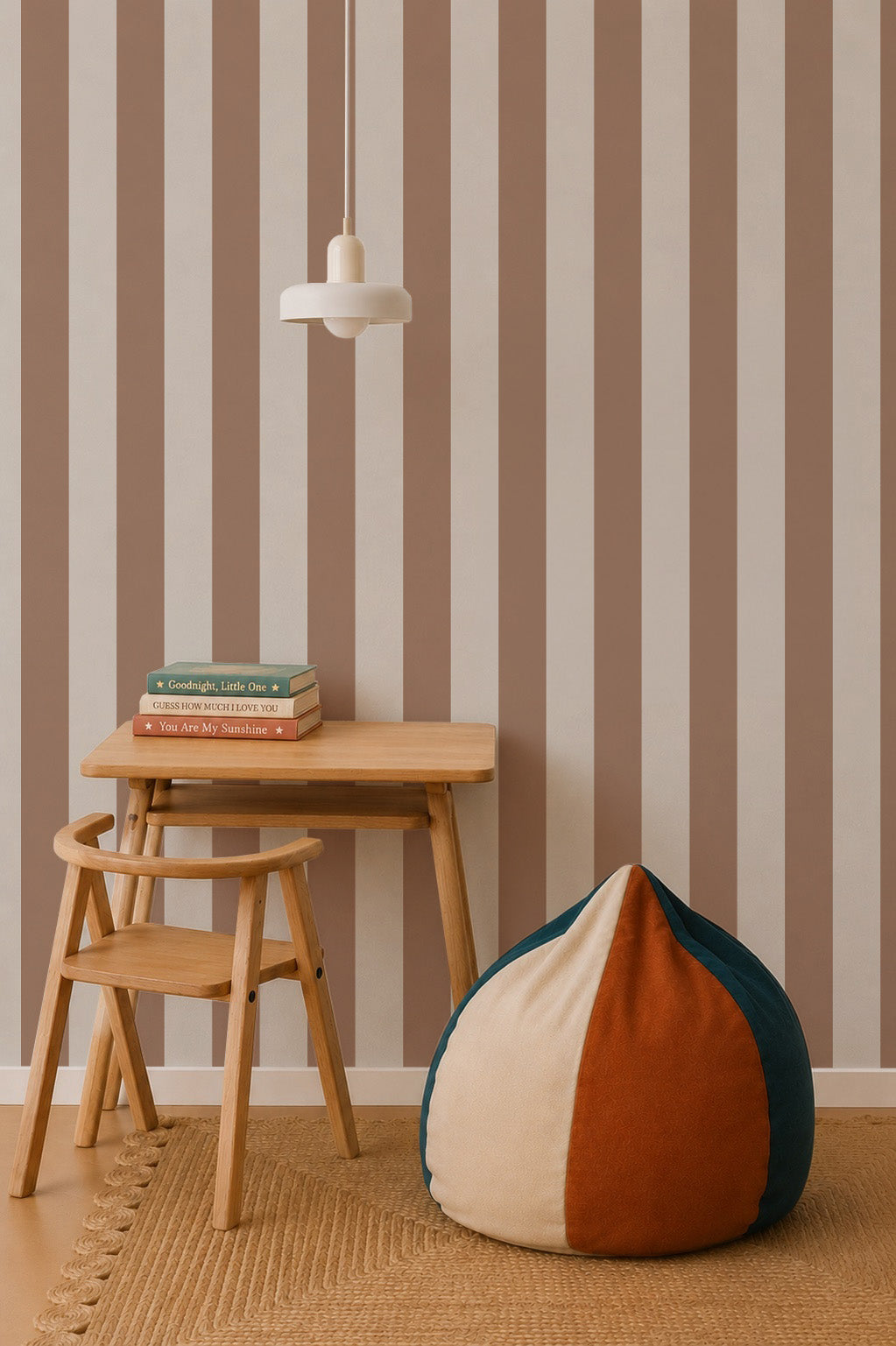 A small wooden desk and chair sit on a woven rug next to a color-blocked bean bag. Three books are stacked under a white pendant light, with vertical Circus Stripe Wallpaper, Latte in beige and brown as the background.