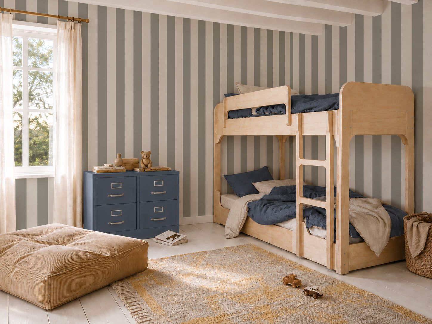 A children’s bedroom features wood bunk beds, a blue dresser, and a tan ottoman, with toys scattered across the rug. Sunlight pours through sheer-curtained windows, illuminating the Circus Stripe Wallpaper in Matcha that introduces playful energy to the space. The combination of neutral and blue tones with vibrant Matcha wallpaper stripes creates a cozy and inviting atmosphere.
