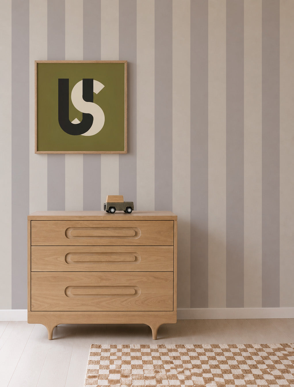 A wooden dresser with three drawers sits against a wall covered in Circus Stripe Wallpaper, Stone, featuring vertical gray and white stripes. A framed abstract artwork and a small toy car are on top, and a checkered rug is on the light wood floor.