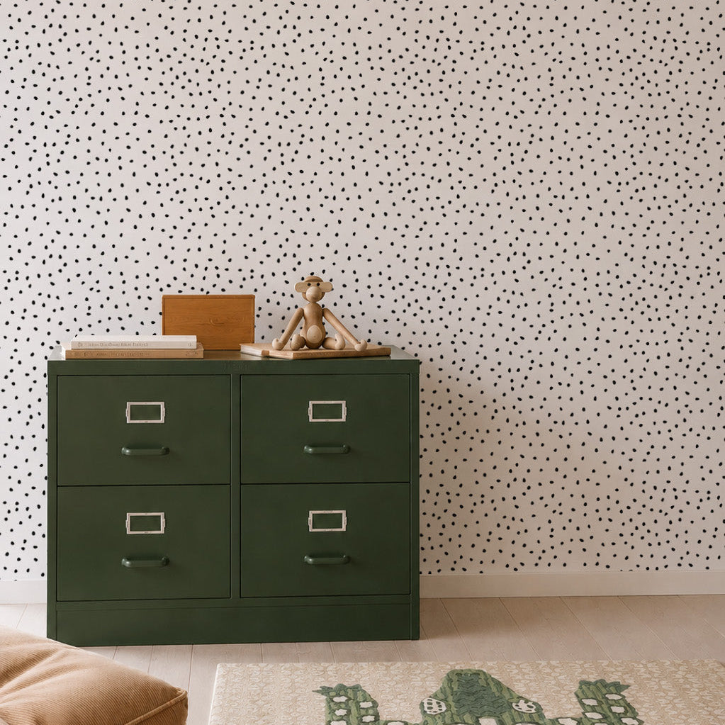 A green four-drawer dresser with a teddy bear and books on top sits against a white wall adorned with the Cora Spotty Wallpaper Sample in Black, adding a playful touch. A beige cushion and a rug featuring a green crocodile design are arranged on the light wooden floor.