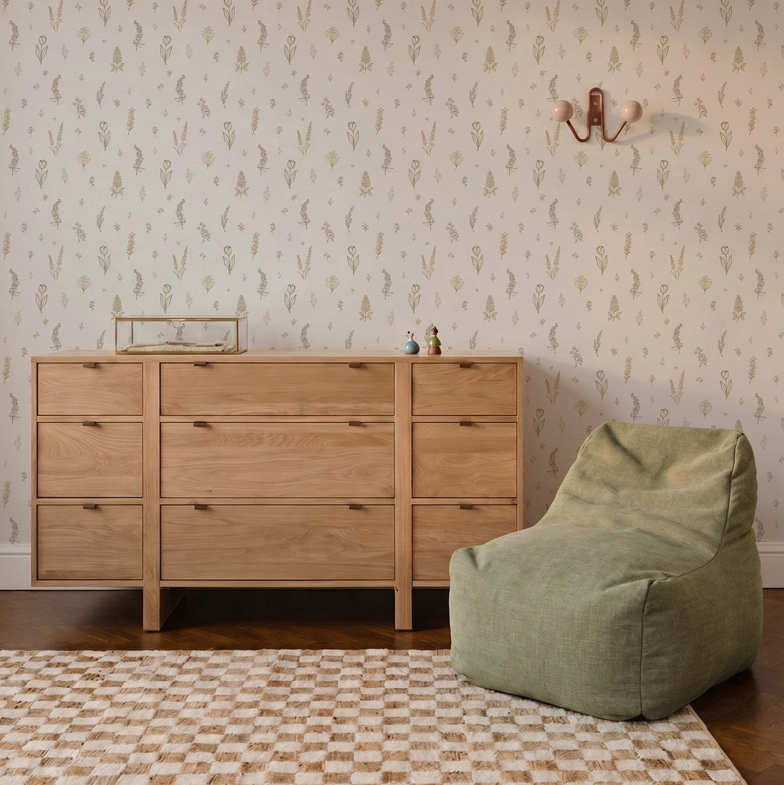 A wooden dresser and a green bean bag chair sit on a checkered rug in front of a wall adorned with Delicate Meadows Wallpaper. A sconce and small decorative items are thoughtfully placed on the wall and dresser.