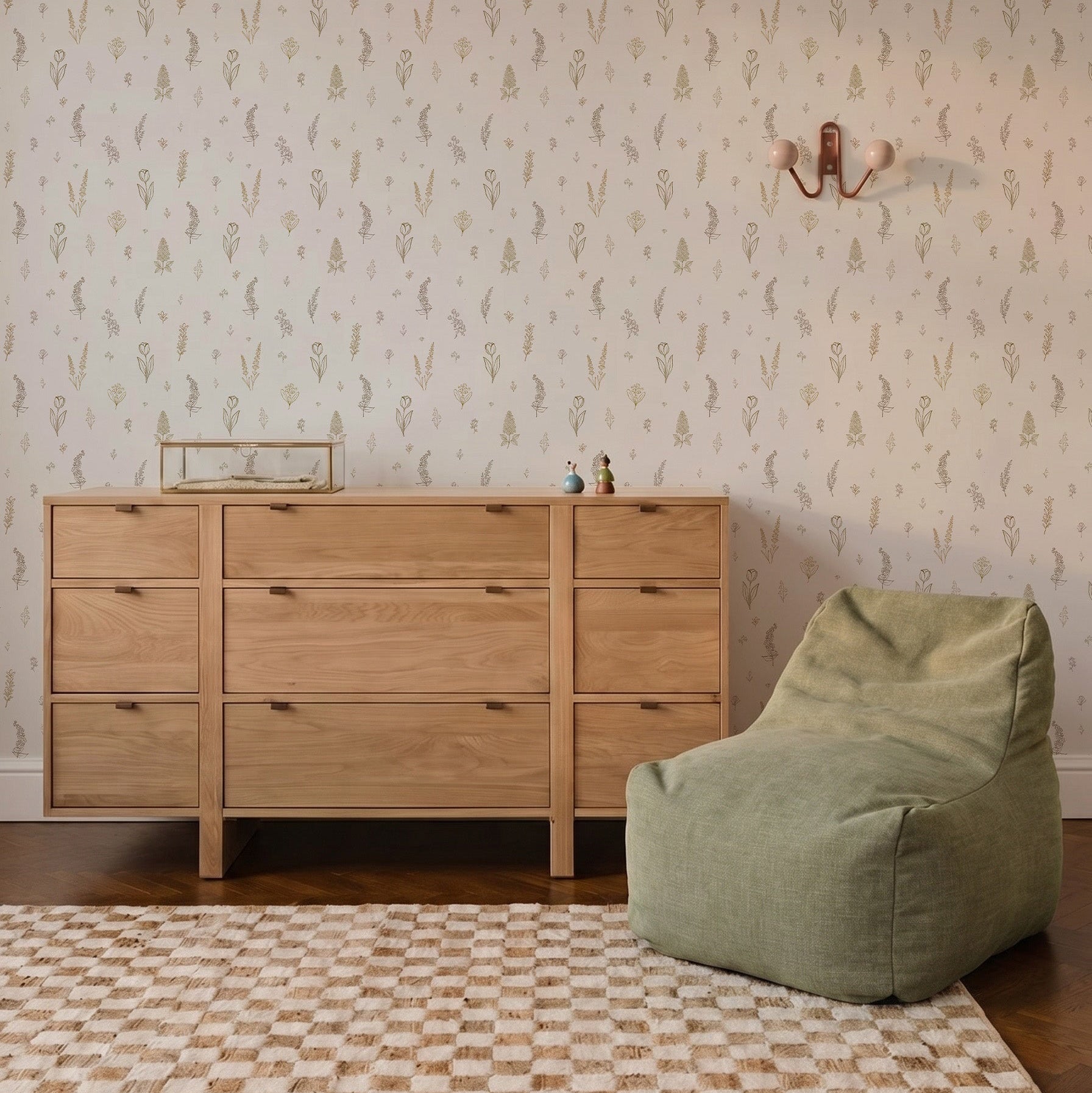 A wooden dresser and a green bean bag chair sit on a checkered rug in front of a wall adorned with Delicate Meadows Wallpaper. A sconce and small decorative items are thoughtfully placed on the wall and dresser.