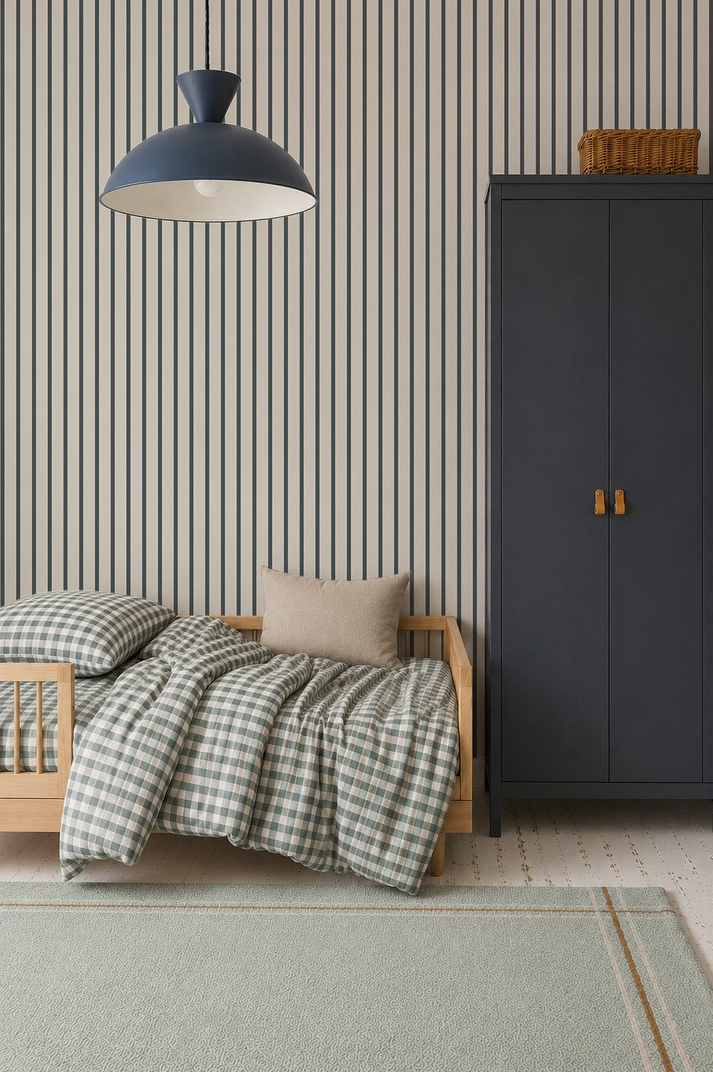 A cozy bedroom features a light wood toddler bed with checkered bedding and a beige pillow. A dark blue wardrobe with a basket on top stands nearby, set against Pinstripe Wallpaper Denim. A large light blue rug covers the floor.