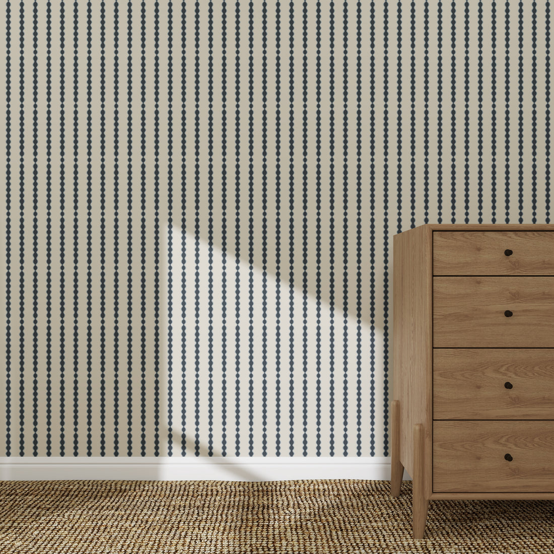 A wooden dresser with four drawers rests on a textured rug in front of a wall decorated with Little Abacus Stripe Wallpaper Sample, Denim. Sunlight casts a bright diagonal shape on the navy and white striped wall.