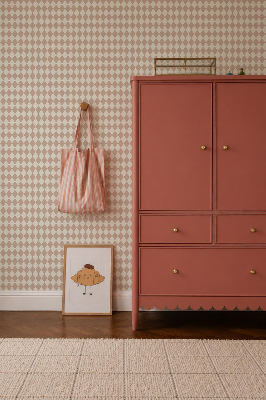 A pink dresser stands on a wooden floor beside a framed croissant drawing, a striped pink bag on a wall hook, and Diamond Dancer Wallpaper in Rose with soft beige harlequin diamond patterns in the background.