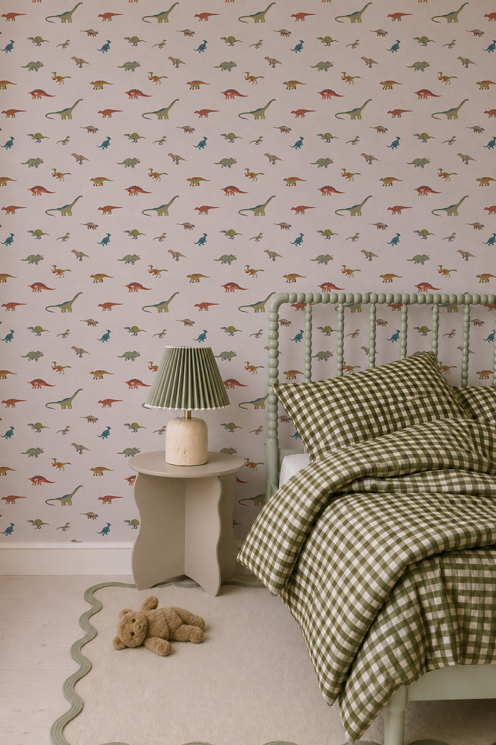A cozy bedroom for little explorers, featuring Dino Discovery Wallpaper, green checkered bedding, a green bed frame, pleated bedside lamp, beige scalloped rug, and a teddy bear.