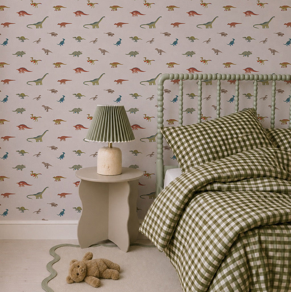 A cozy bedroom for little explorers, featuring Dino Discovery Wallpaper, green checkered bedding, a green bed frame, pleated bedside lamp, beige scalloped rug, and a teddy bear.
