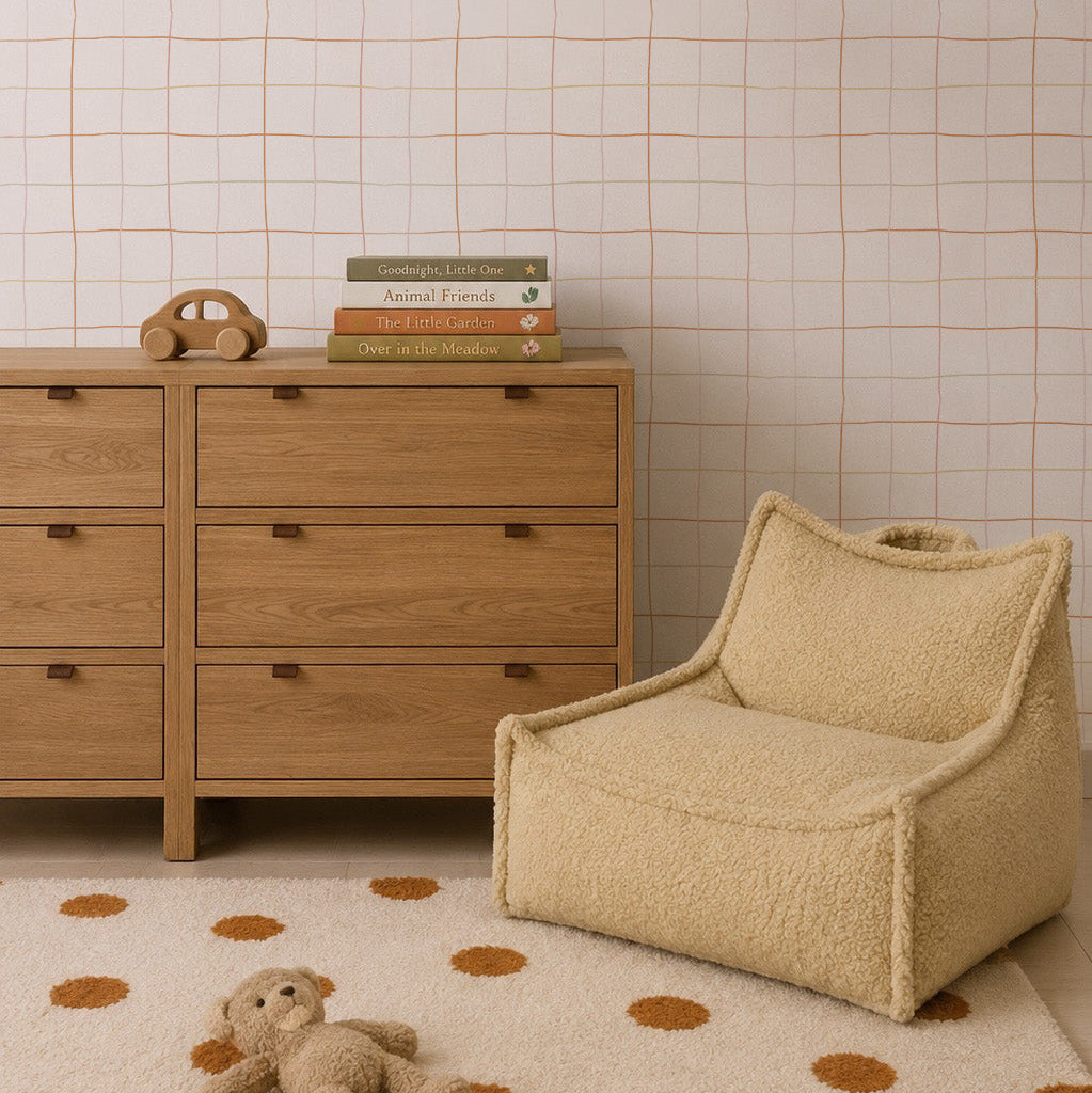 Minimalist nursery with a wooden dresser, children’s books, toy phone, beige fuzzy chair, polka dot rug featuring a plush teddy bear, and walls adorned with Doodle Grid Wallpaper—a subtle repeat pattern ideal for modern interiors.
