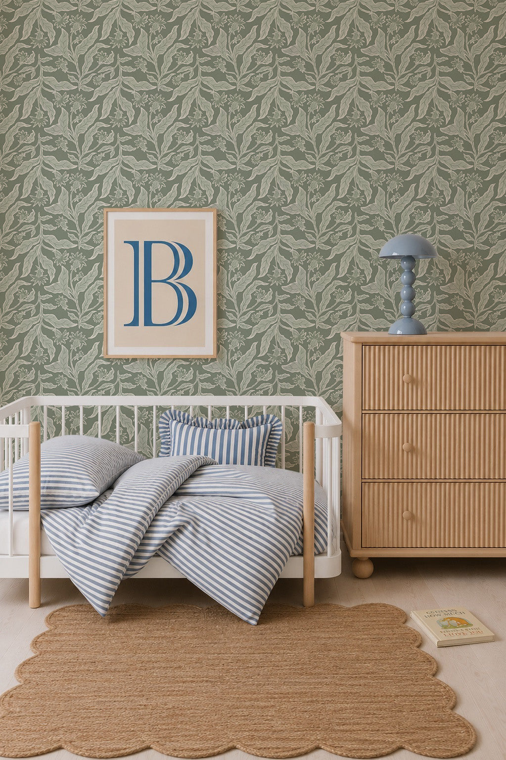 A cozy nursery features a white crib with blue striped bedding, a wooden dresser with a blue lamp, a scalloped brown rug, Elodies Thistle Wallpaper in sage, and a framed letter B on the wall.