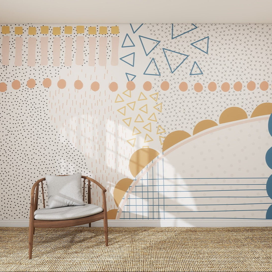 A modern room with a wooden chair and cushion sits before the Evie Abstract Wallpaper Mural in blue, mustard yellow, and pink—featuring triangles, dots, lines, and arches—perfect as colourful children’s room wallpaper above a woven rug.