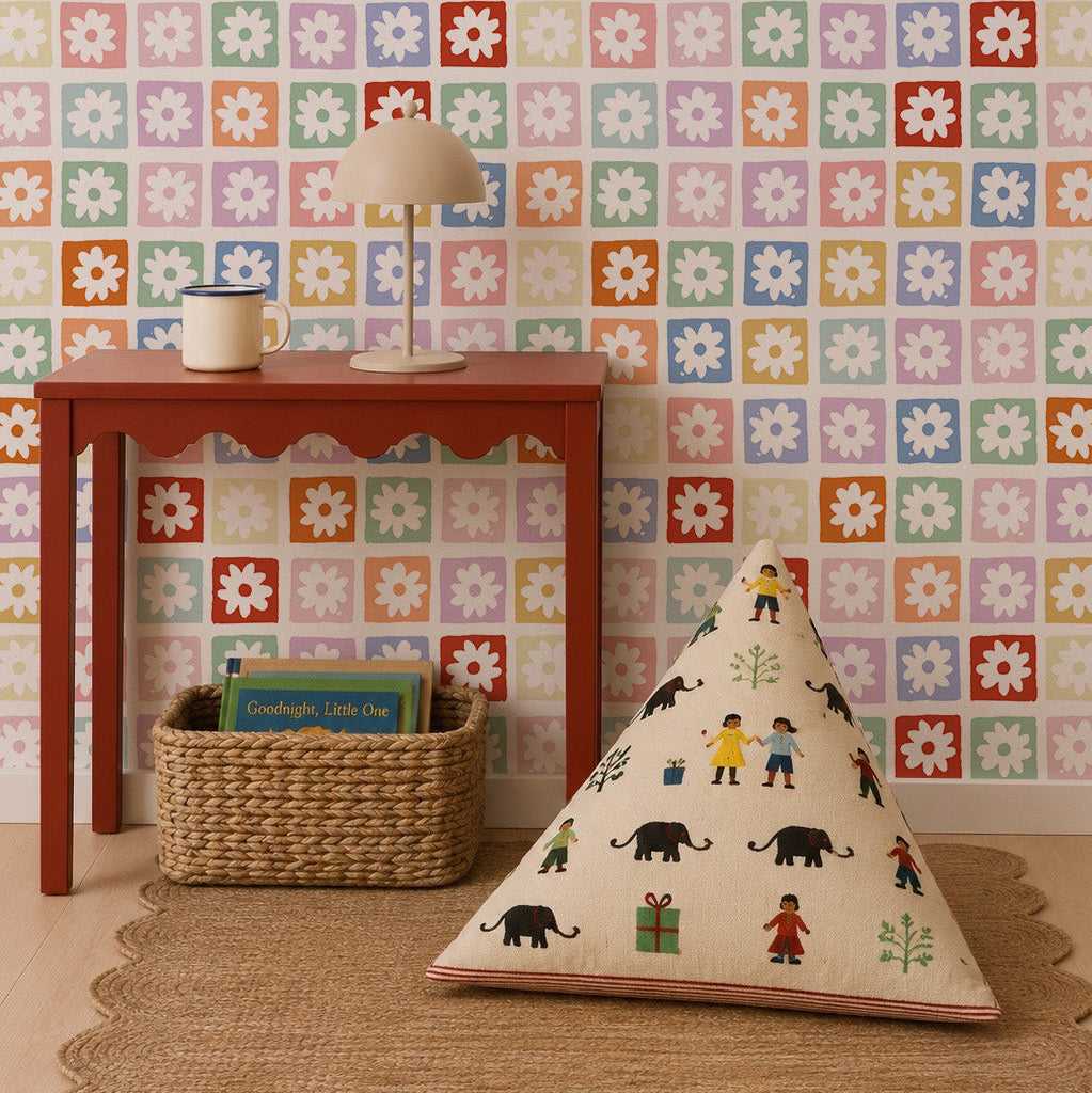 A colorful room features a red table with a lamp and cup, a woven basket with books underneath, a patterned floor cushion, and walls adorned in the Floral Funland Mural showcasing multicolored floral squares.