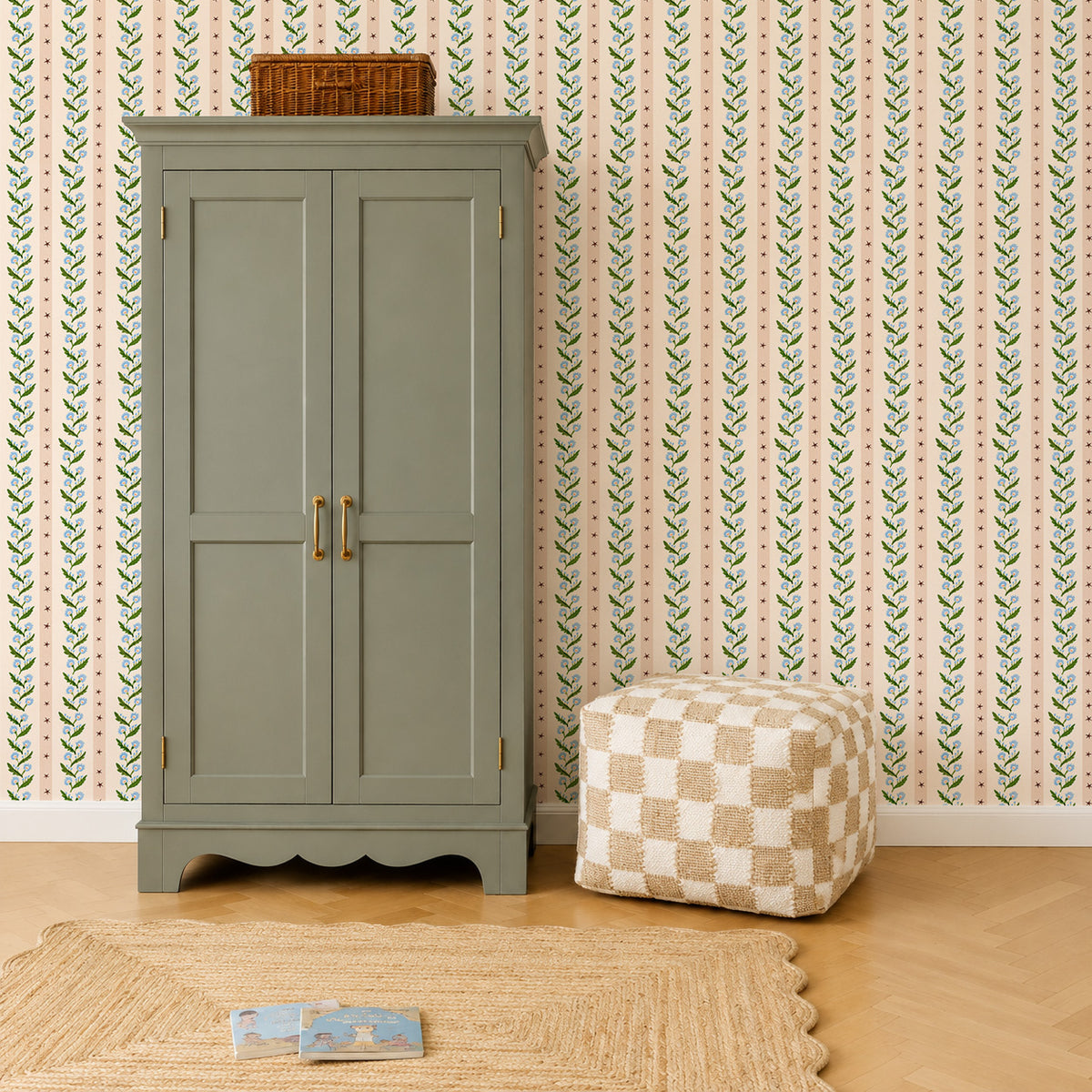 A sage green wardrobe stands against Forget-Me-Not Trail Stripe wallpaper featuring vertical blue flowers and dark green vines, with a beige checkered ottoman and a woven rug on the floor.