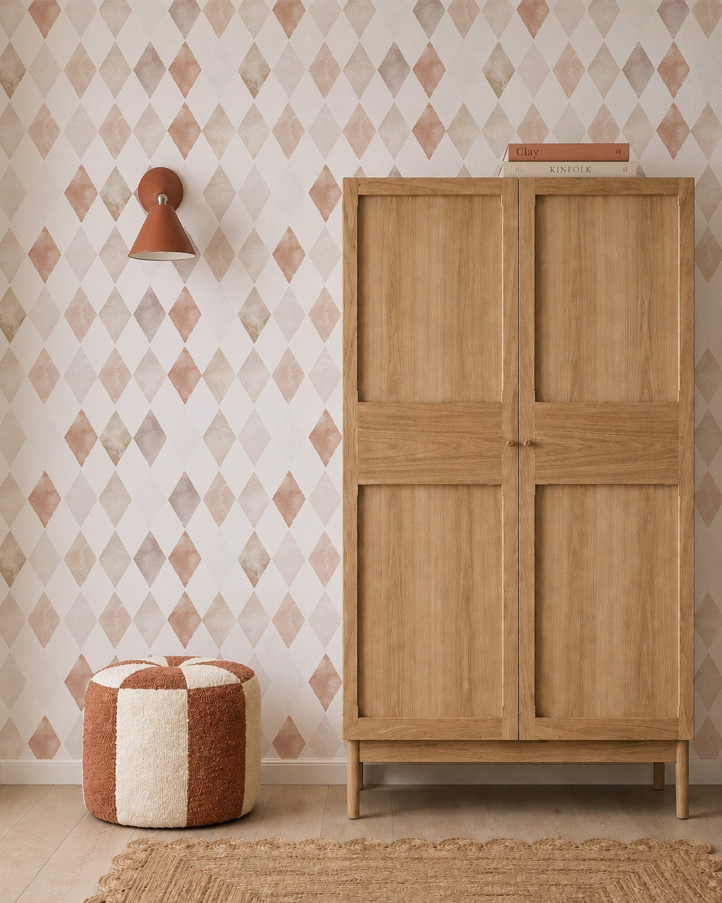 A wooden cabinet stands against Harlequin Watercolour Wallpaper, Pink. To the left is a round knit pouf in cream and rust, with a small wall sconce above. A book and small item sit atop the cabinet.
