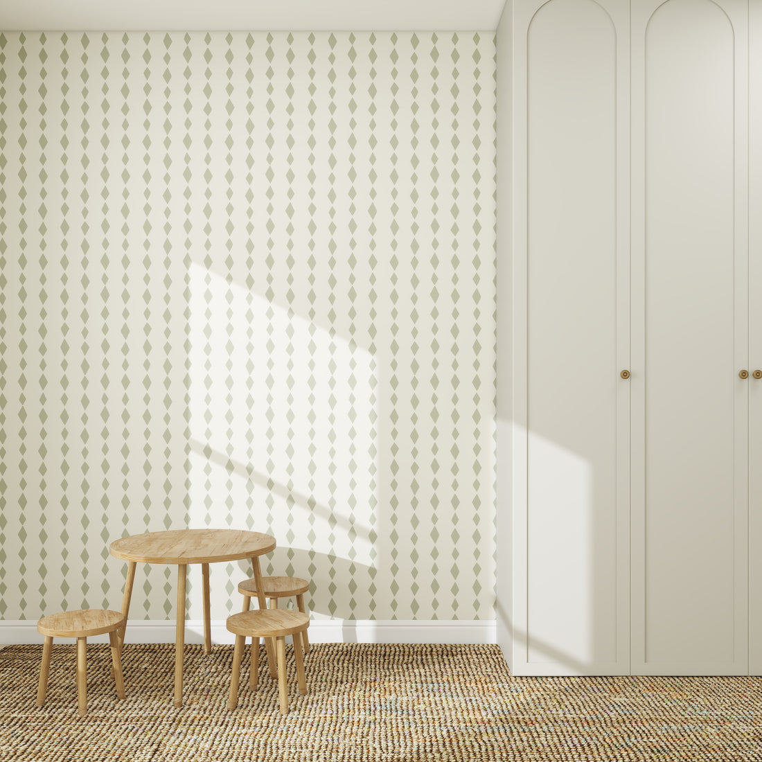 A kids’ room with a small wooden table, three stools on a woven rug, and patterned Sage Green Jolly Jester Wallpaper with dots. A tall white wardrobe stands nearby as sunlight casts bright shadows across the wall and floor.