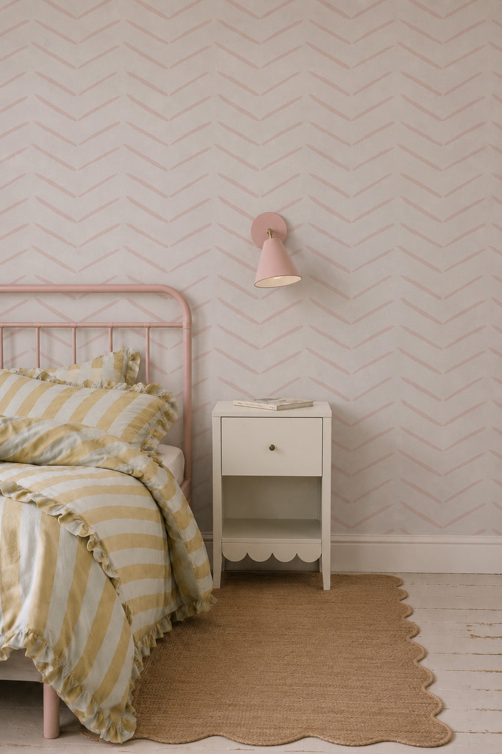 A cozy bedroom features a pink metal bed with yellow and white striped bedding, a scalloped-edge nightstand, a book, a pink wall lamp, rose accents, Herringbone Wallpaper, Rose, and a woven rug on white wood floors.