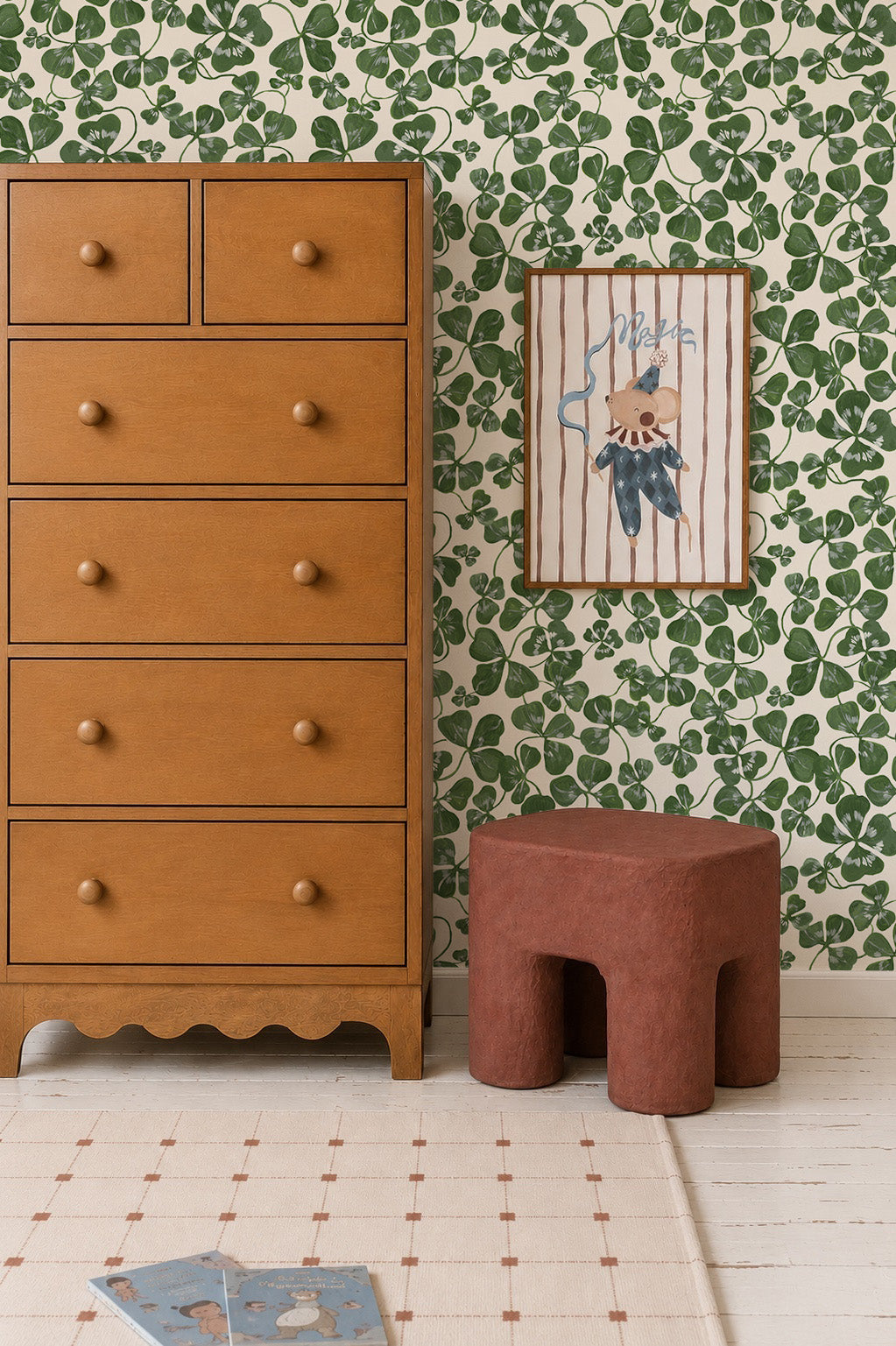 A cozy room with a tall wooden dresser, a brown textured stool, a framed bear illustration on How Lucky We Are Clovers Wallpaper, plus a light rug and book on a white floor.