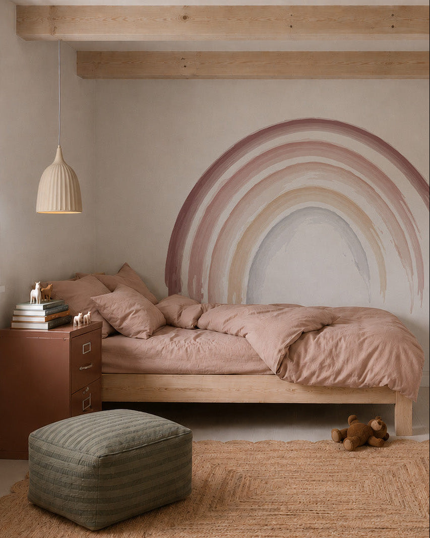 A cozy bedroom with a wooden bed, pink bedding, pouf, woven rug, and teddy bear. An Iris Watercolour Rainbow Mural Sample decorates the wall above a brown cabinet with toy animals; a pendant light hangs overhead.