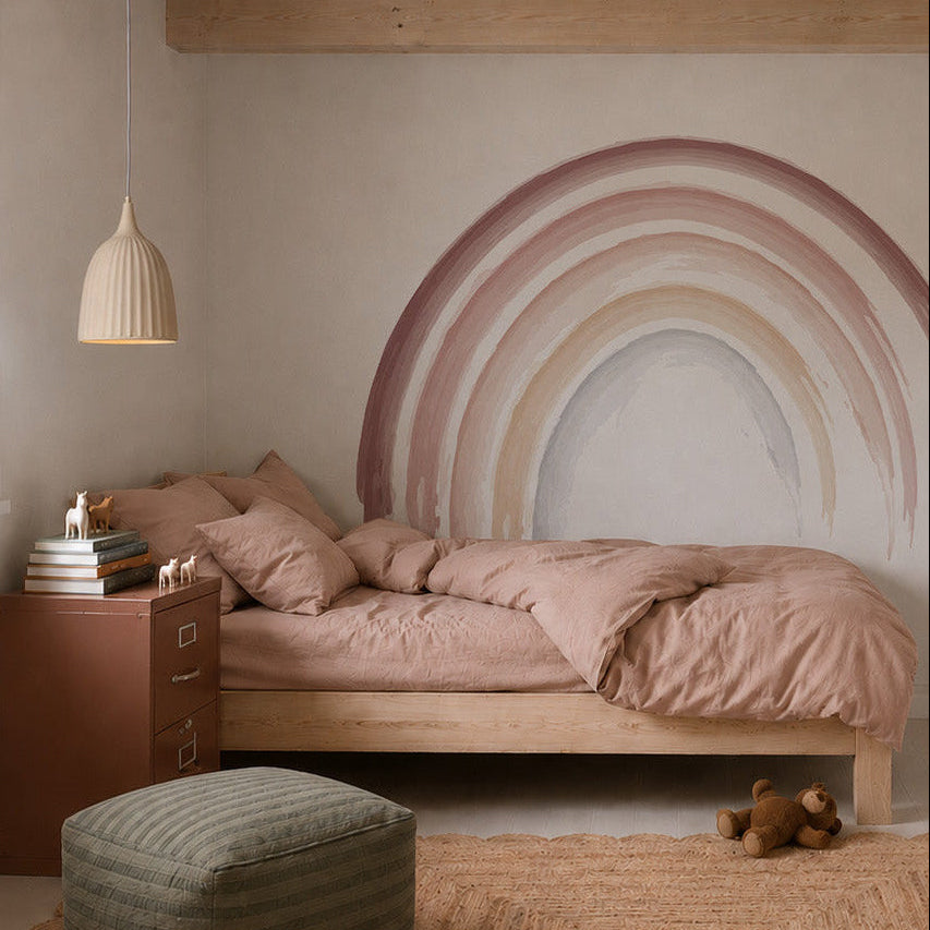 A cozy bedroom with a wooden bed, pink bedding, pouf, woven rug, and teddy bear. An Iris Watercolour Rainbow Mural Sample decorates the wall above a brown cabinet with toy animals; a pendant light hangs overhead.