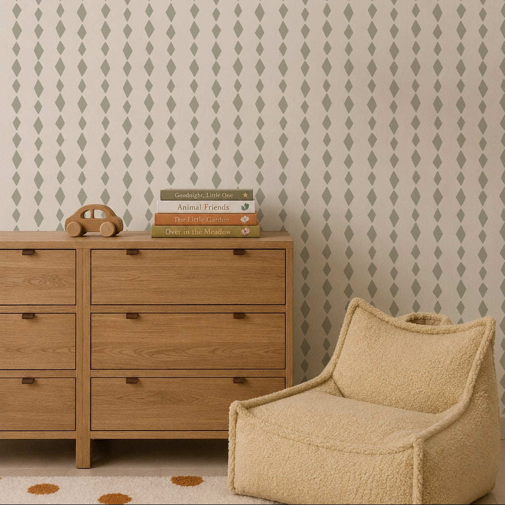 A neutral-toned room with a wooden dresser, beige textured armchair, and stacked books. The wall features Jolly Jester Wallpaper in sage green, adding playful elegance perfect for kids&