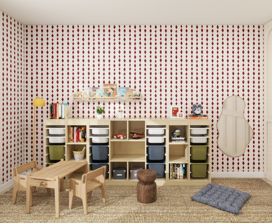 A children’s playroom features Jolly Jester Wallpaper Repeat Cherry for a pop of color, cubby storage with books and toys, a small wooden table and chairs, a floor cushion, quirky mirror, large cabinets, and natural light from the window.
