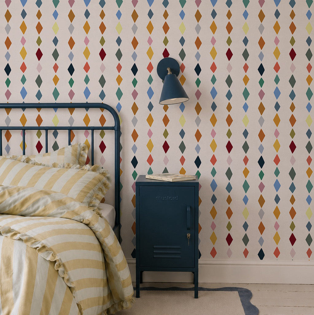 A cozy bedroom features a metal bed frame, yellow and white striped bedding, blue nightstand, wall lamp, painted wooden floorboards with a rug, and walls adorned with Jolly Jester Wallpaper, Multi—colorful diamonds in vertical rows.