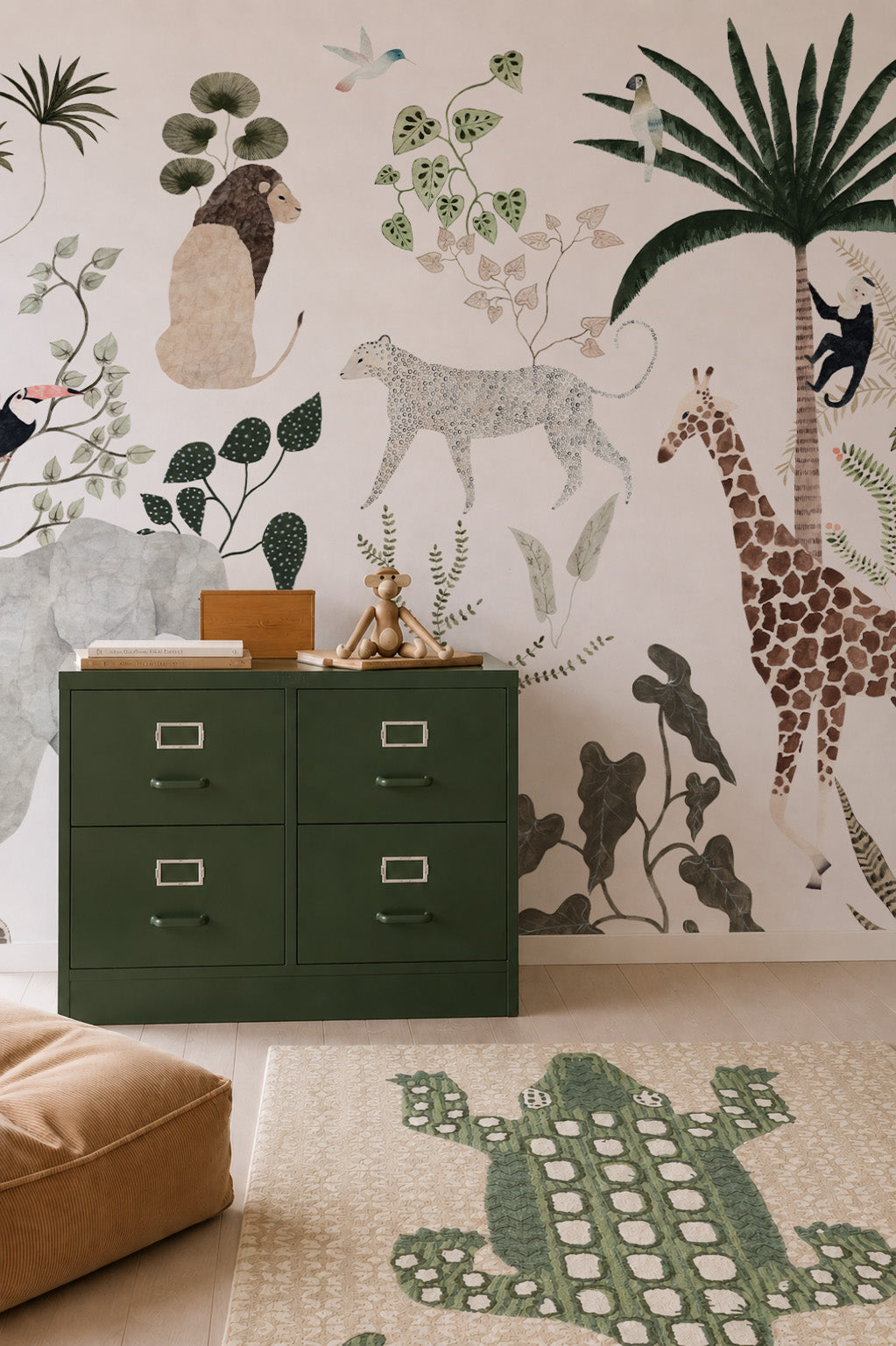 A green filing cabinet, plush toy, and brown cushion in a room decorated with the Jungle Dreams Mural—kids’ wallpaper featuring animals like a giraffe, monkey, and bird—and a crocodile-patterned rug on a light wood floor.