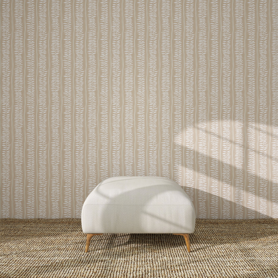 A white upholstered ottoman with wooden legs rests on a textured brown rug before Meadow Leaf Stripe Wallpaper in Clay, as sunlight highlights the botanical charm of the leaf-patterned backdrop.