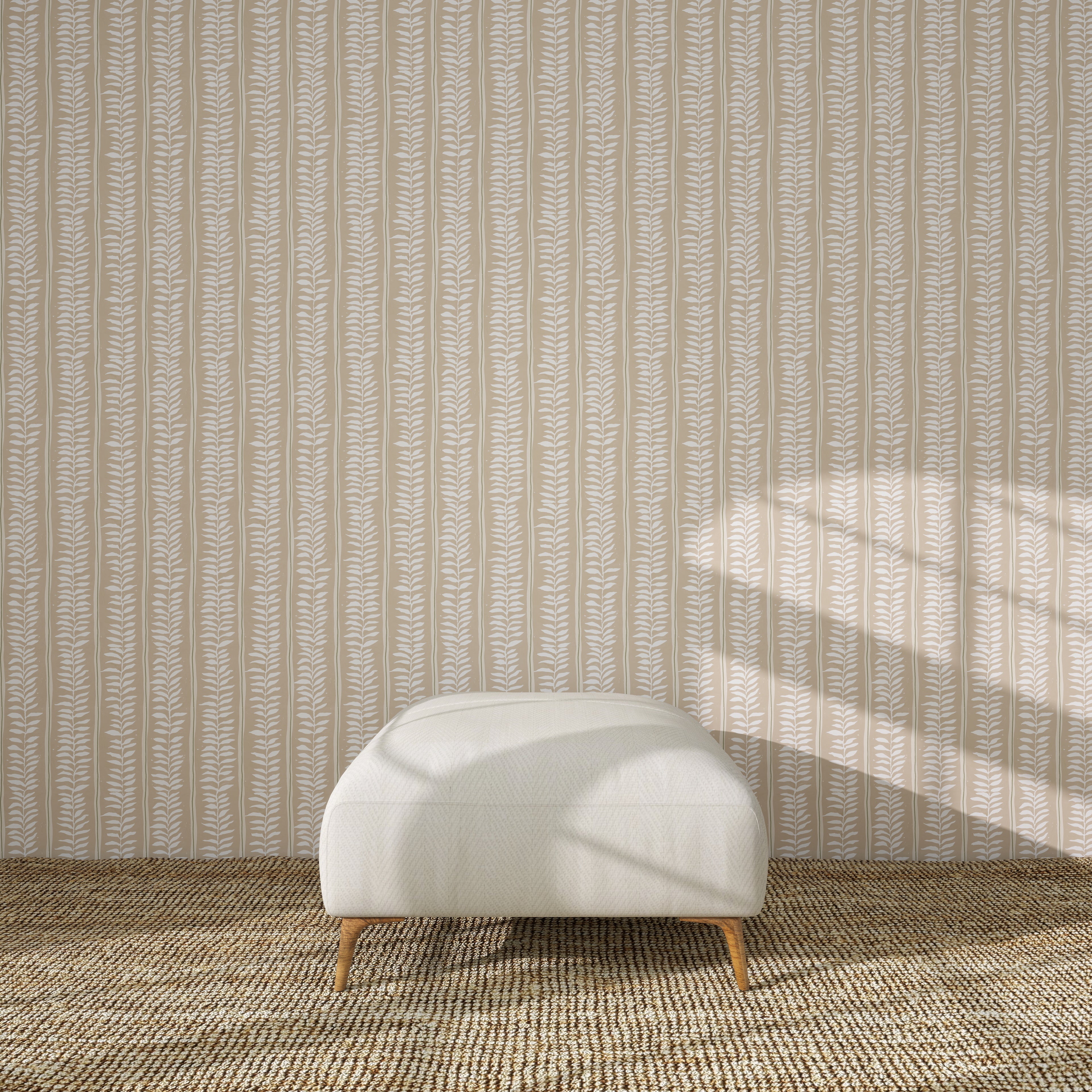 A white upholstered ottoman with wooden legs rests on a textured brown rug before Meadow Leaf Stripe Wallpaper in Clay, as sunlight highlights the botanical charm of the leaf-patterned backdrop.