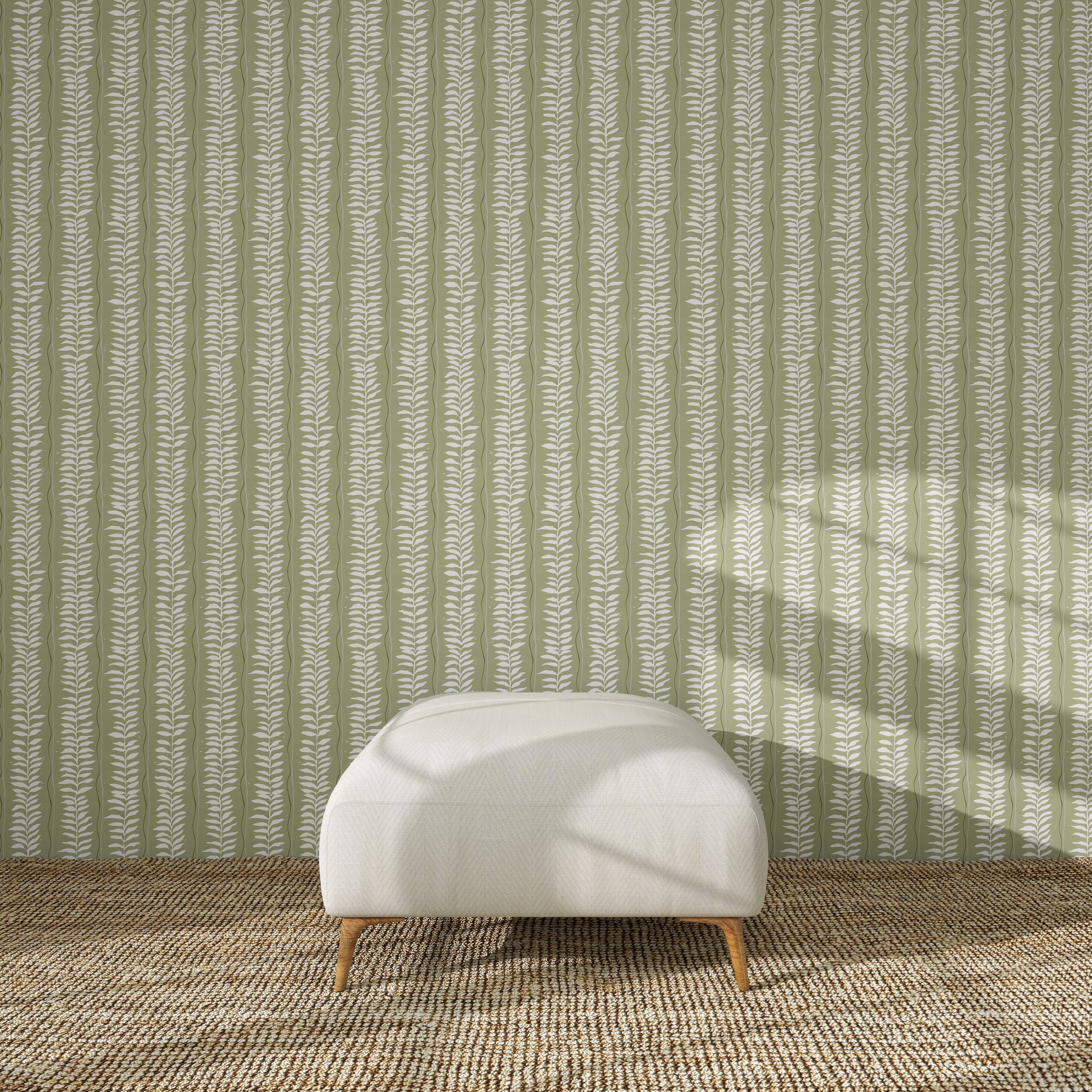 A white upholstered ottoman with wooden legs rests on a textured brown rug, in front of a green wall decorated with the Meadow Leaf Stripe Wallpaper Sample in Sage. Sunlight casts soft shadows across the scene.