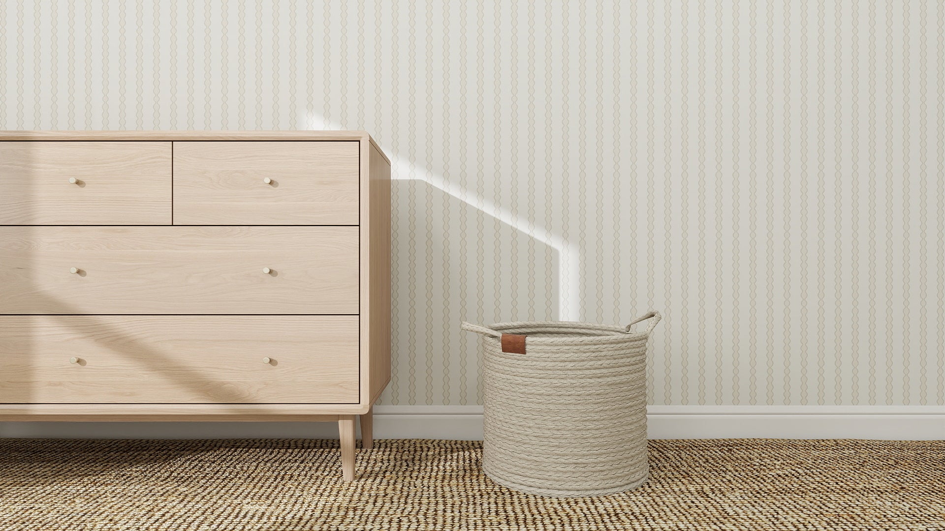 A light wood dresser with four drawers stands on a woven rug in a neutral nursery, next to a textured cream basket and Little Abacus Stripe Wallpaper in Off White and Taupe. Sunlight casts a bright diagonal line across the wall and dresser.
