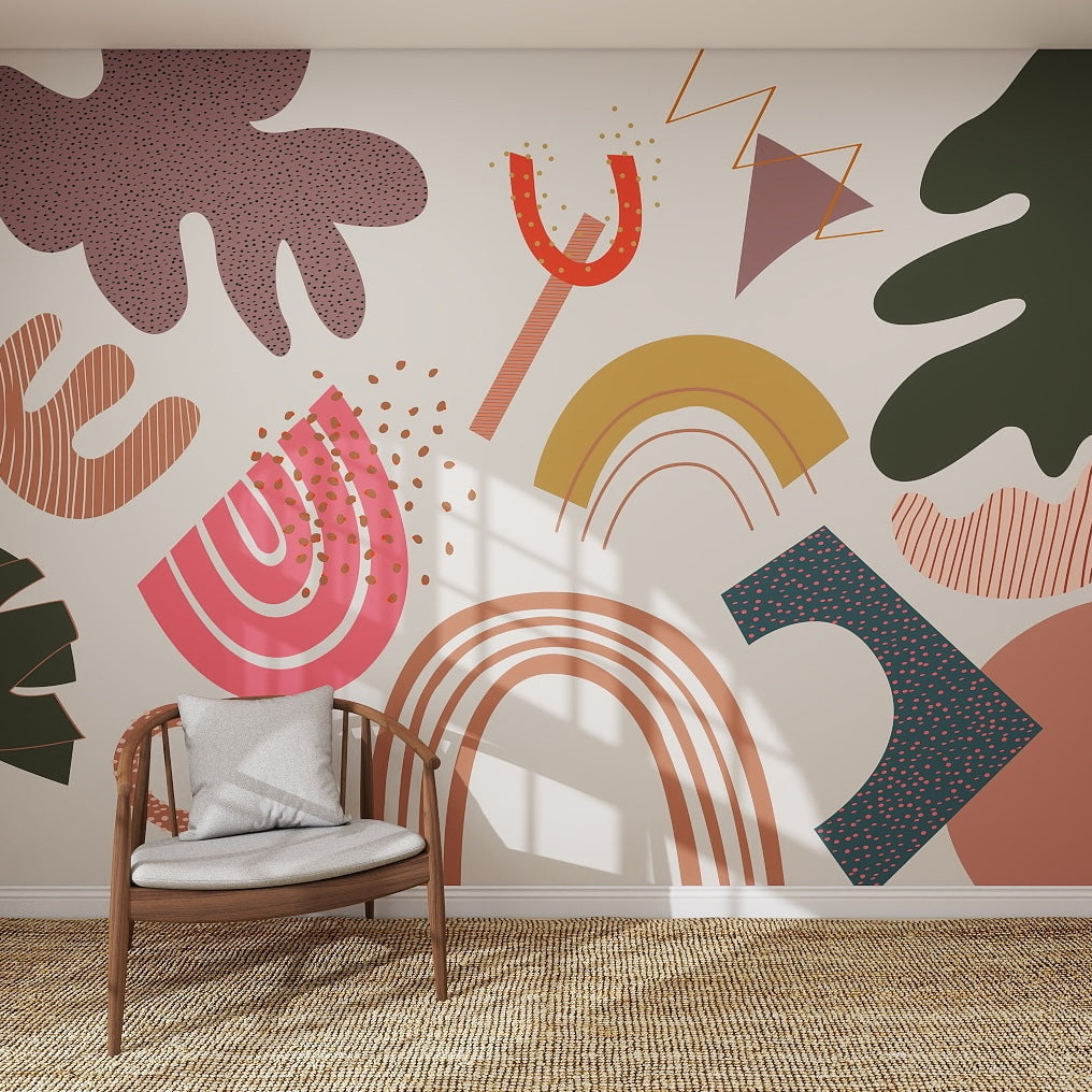 A modern room with a wicker chair and gray cushion faces a white wall decorated with the Little Erin Abstract Wallpaper Mural—colorful shapes like rainbows, leaves, stripes, and dots. Sunlight streams in, highlighting its playful design.