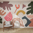 A modern room with a wicker chair and gray cushion faces a white wall decorated with the Little Erin Abstract Wallpaper Mural—colorful shapes like rainbows, leaves, stripes, and dots. Sunlight streams in, highlighting its playful design.