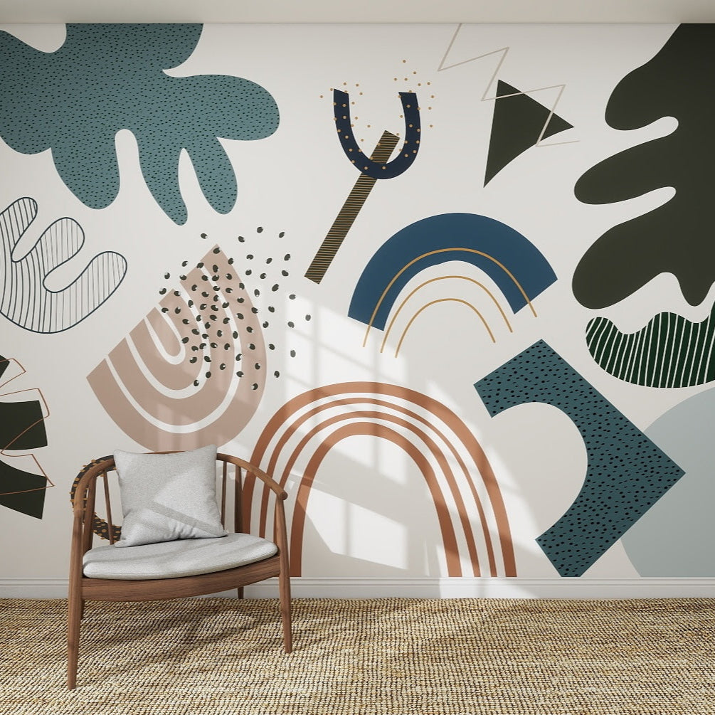 A wooden chair with a gray cushion rests on a woven rug in front of the Little Erin Abstract Wallpaper Mural | Blue, featuring bold blue, green, brown, and beige shapes. Sunlight casts shadows across the vibrant wall pattern.