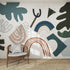A wooden chair with a gray cushion rests on a woven rug in front of the Little Erin Abstract Wallpaper Mural | Blue, featuring bold blue, green, brown, and beige shapes. Sunlight casts shadows across the vibrant wall pattern.