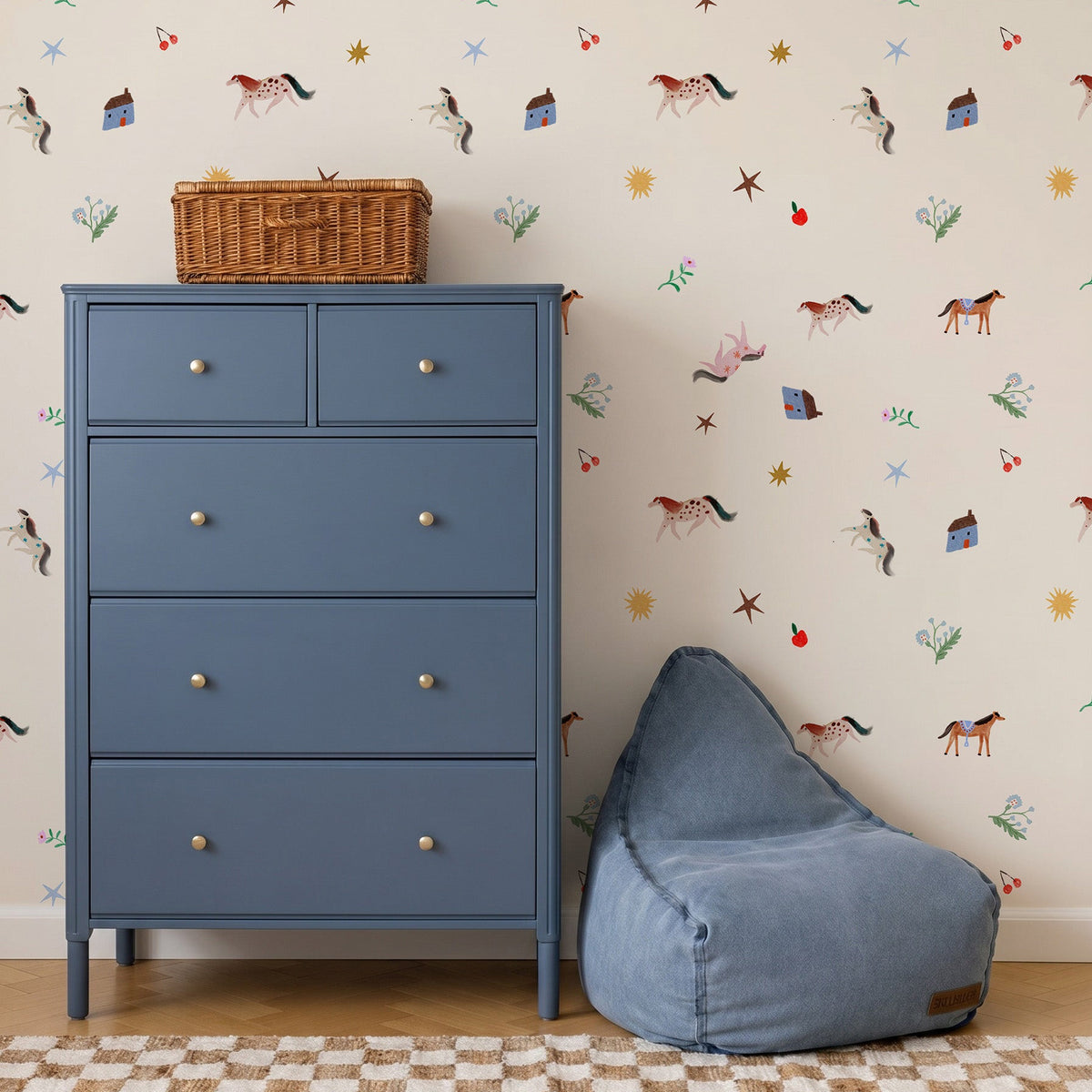 A blue chest of drawers and a bean bag chair sit in front of Little Paddock Wallpaper, which features a repeating pattern of horses, small houses, stars, and flowers.