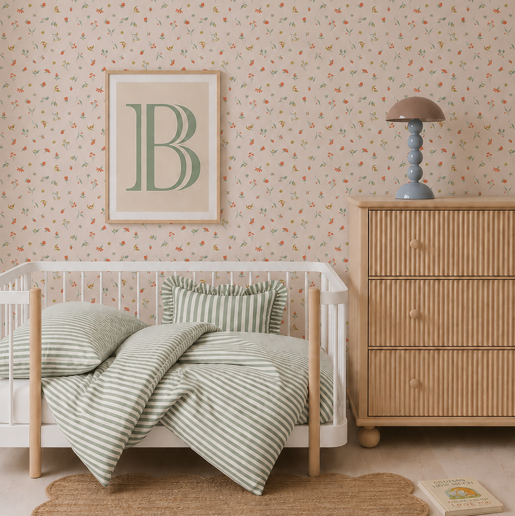 A nursery with a wooden crib, green striped bedding, a rattan dresser topped with a lamp and three stacked books, a rug, and Lucys Spring Buds Wallpaper featuring a delicate repeat pattern. A framed letter B hangs above the crib.
