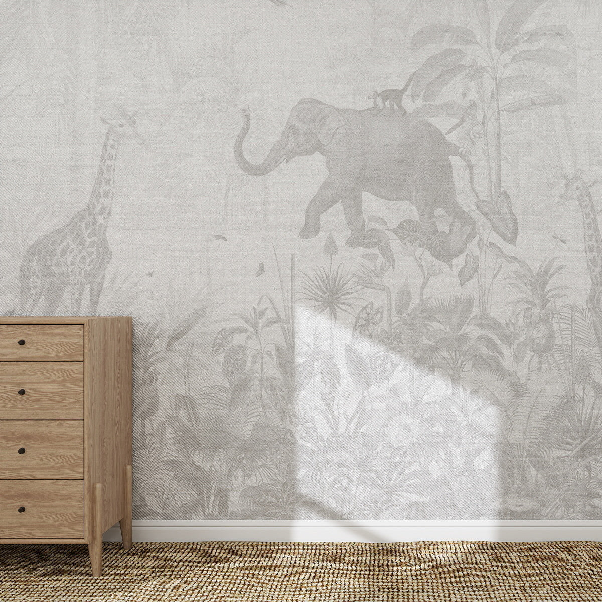 The Magic Jungle Mural, Monochrome decorates the wall with elephants, giraffes, and lush plants. A light wood chest of drawers sits on a woven rug in front, bathed in sunlight across this tranquil animal-themed wallpaper.