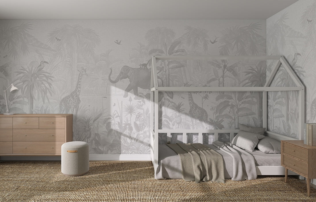 A children’s bedroom with a house-shaped white bed, neutral bedding, wooden dresser, round pouf, and a small nightstand. The Magic Jungle Monochrome Wallpaper Mural with giraffes and elephants adds a cozy touch to the kids room.