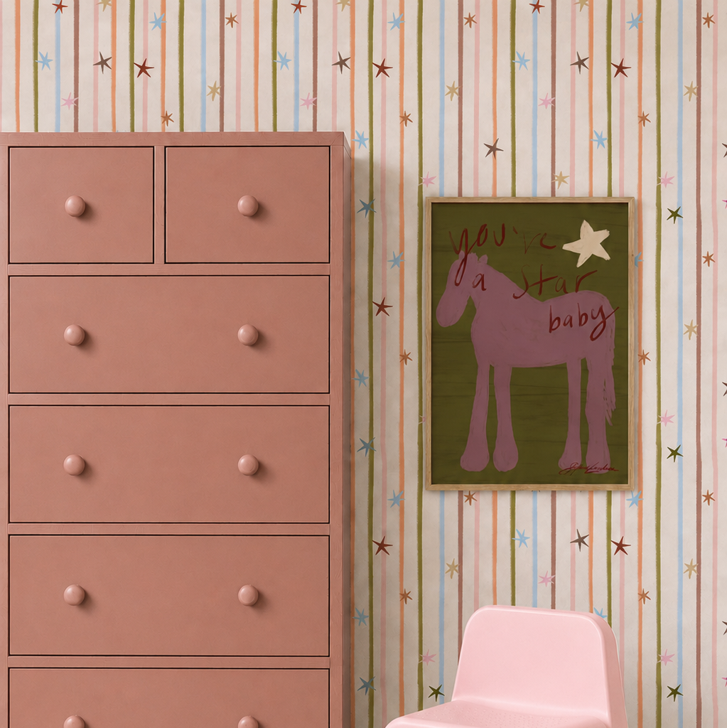 A dusty pink dresser stands against Starlight Stripe Wallpaper, featuring vertical multicolored stripes and small stars, with a framed horse illustration hung on the wall.