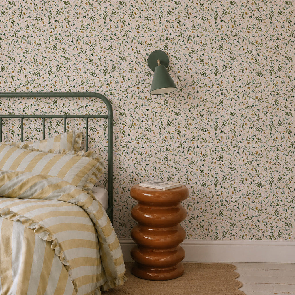 A cozy bedroom with a green metal bed, yellow and white striped bedding, a brown sculptural side table, a tan woven rug, and Meadow Flutter Leaves Wallpaper with hand-painted leaves, plus a green wall lamp above the table.