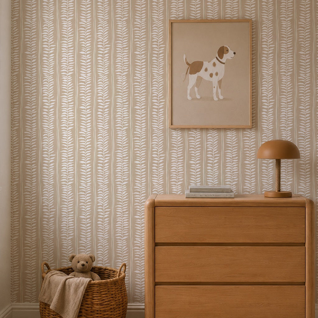 A cozy room features a light wood dresser, brown lamp, books, and a framed dog illustration above. By the dresser sits a woven basket with a teddy bear and blanket. The wall is adorned with Meadow Leaf Stripe Wallpaper in Clay.