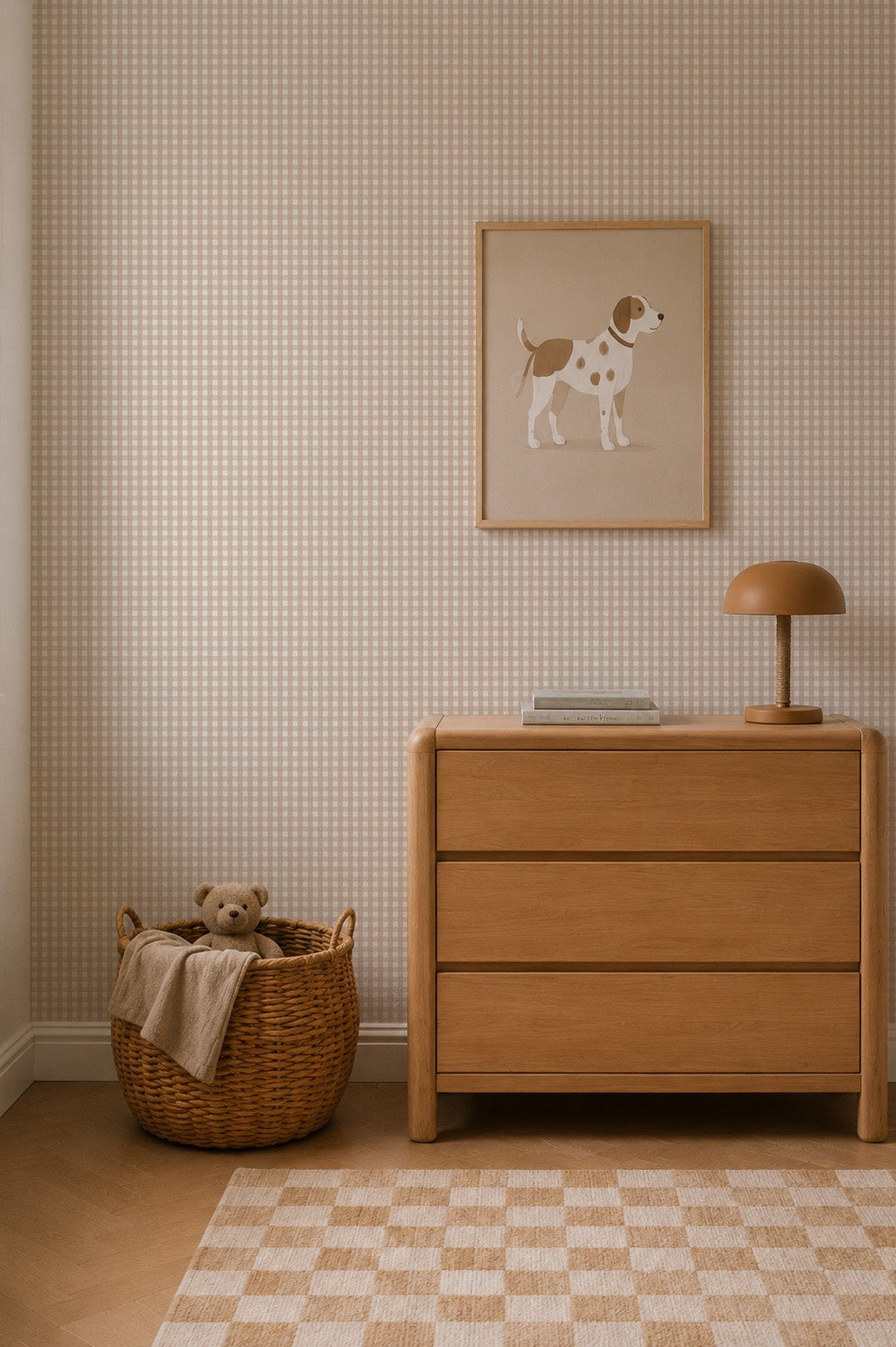 A cozy nursery features a wicker basket with a teddy bear, a wooden dresser with books and a lamp, and a framed dog illustration hanging above against the charming Mini Check Wallpaper in Pink.