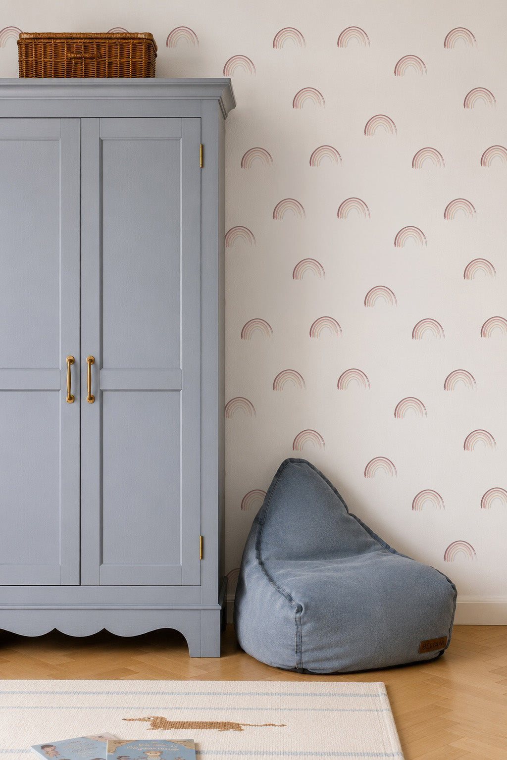 A light blue armoire with gold handles stands next to a blue bean bag chair in a room with white walls decorated with Mini Watercolour Rainbow Wallpaper. A woven basket atop the armoire complements the wallpaper&