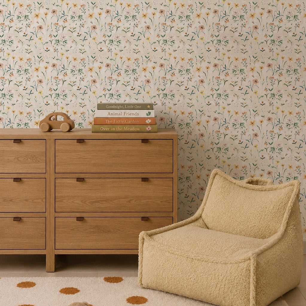 A cozy room with a wooden dresser, stacked books, a toy phone, a fluffy beige chair, and hand-painted Morning Meadow Wallpaper featuring wildflowers. Brown polka dots are on the carpet.