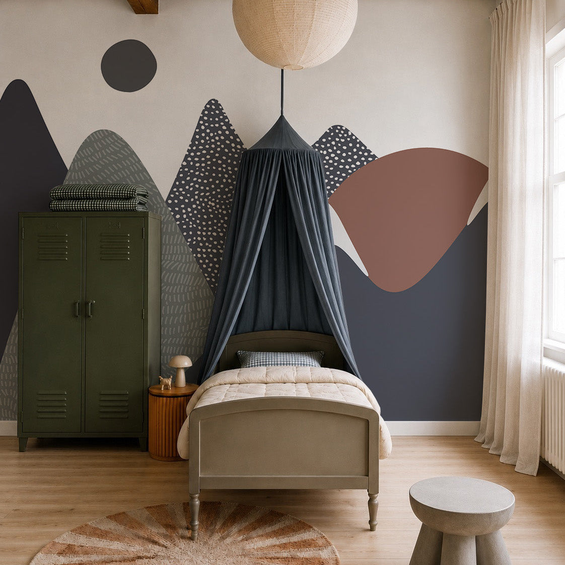 A cozy kids’ bedroom features a single bed beneath a dark canopy, round ceiling light, green locker wardrobe, stacked blankets, sunburst rug, and the Mountain Mural as a charming focal point for nature-inspired décor.